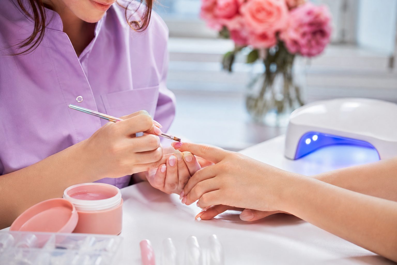 Technician applying builder gel to a client's nail beside soft gel tips and a curing lamp